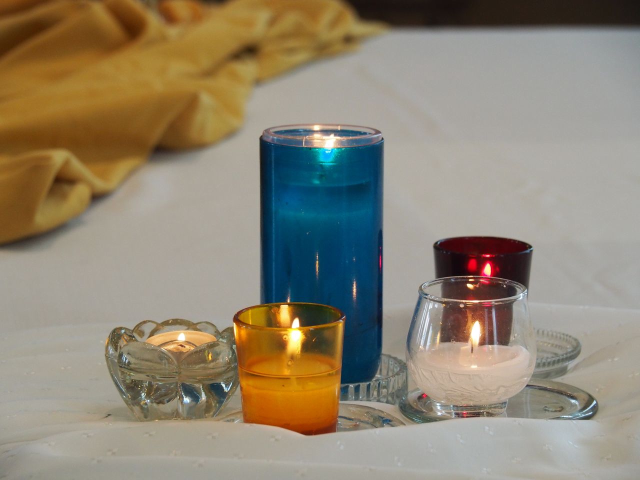 Home Five colorful candles lit on white linen, creating a warm and serene atmosphere.